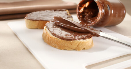 Bread slice with chocolate hazelnut spread or butter. Spreading hazelnut cream, chocolate nut butter with knife. Slow motion. Sunny morning. Traditional breakfast. Angle view. 