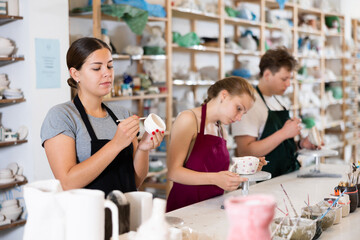Young woman teacher in apron paints ceramic cup in ceramic workshop..