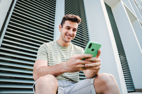Young guy with friendly expression smiling and having fun playing a video game with his cellphone sitting outdoors. Caucasian teenage stundent enjoying using a mobile phone for watch the social media