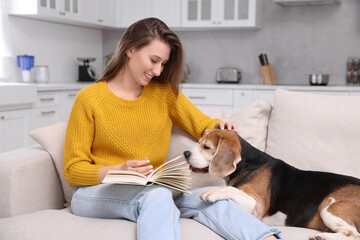 Happy young woman reading book near her cute Beagle dog on couch at home. Lovely pet