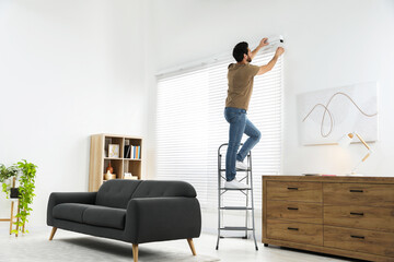 Man on metal folding ladder installing blinds at home. Space for text