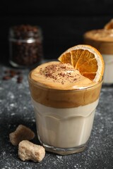 Glass of delicious dalgona coffee with dry orange, sugar and chocolate on grey textured table