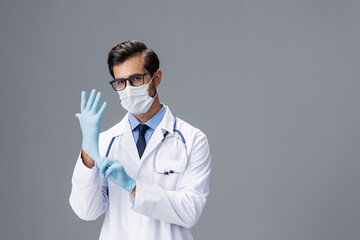 Male doctor in a white coat and medical mask and sterile gloves, eyeglasses looks at the camera on a gray isolated background, copy space, space for text, health