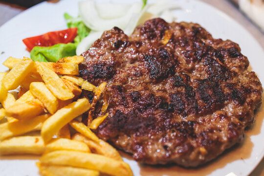 Pljeskavica with french fries on a plate, traditional serbian dish, grilled dish of spiced meat patty mixture of pork, beef and lamb, popular balkan steet food Pleskavitza, served in cafe
