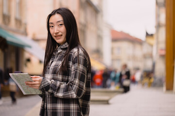 Portrait of a beautiful brunette Korean woman holding a map and comparing it with smartphone navigation on the streets of the old city. Asian tourist woman traveling in Europe.