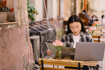 Portrait of a beautiful brunette Korean woman sitting in an authentic cafe and using a laptop, choosing travel routes on a map in an old European city.