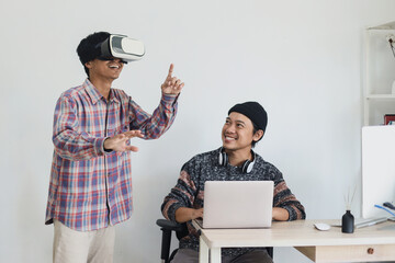 Two young Asian friends wearing virtual reality VR goggles headset and holding smartphone as joystick game console, having fun playing online 3D simulator game