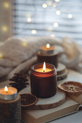 Cozy autumn or winter composition with aromatic candle, wool sweater, fairy lights, book. Aromatherapy, home atmosphere of cosiness and relax. Wooden background close up