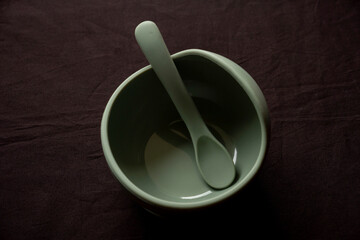 Empty dishes for baby, soft shatterproof tableware for the first feeding, spoon and bowl for baby food on black texture background.