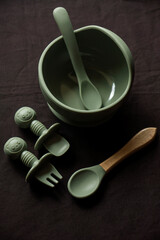 Empty dishes for baby, soft shatterproof tableware for the first feeding, fork, spoons and bowl for baby food on black texture background.