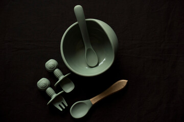 Empty dishes for baby, soft shatterproof tableware for the first feeding, fork, spoons and bowl for baby food on black texture background.