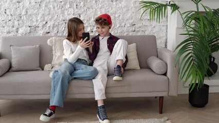 Teenagers boy and girl are sitting on a sofa in a loft-style bright room. Friends listen to music, podcasts from smartphone through headphones. One pair of headphones. Teenage relationships. Video 4K - Powered by Adobe