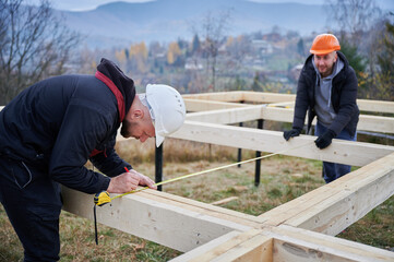 Man worker building wooden frame house on pile foundation. Carpenter using tape measure for...