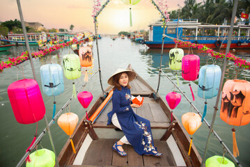 Asian woman is wearing Ao Dai traditional Vietnamese dress and sitting on a lantern boat ride on the river at Hoi An old town in Vietnam.