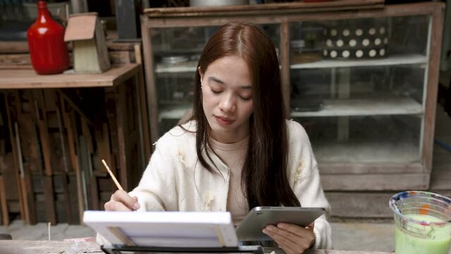 Cute Asian woman art student looks at the sample picture on the tablet and then practice drawing accordingly. focus on painting Practicing free time during the day. Use a brush to paint on white cloth