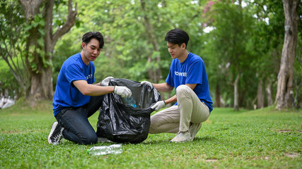 Two Asian men with garbage bag helping each other clean up the area in a public park.