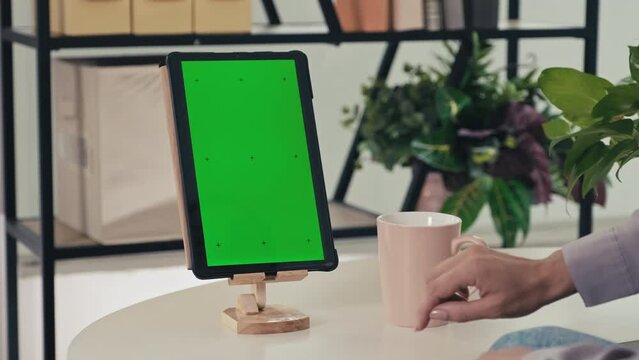 Close-up shot of womans finger swiping up green chroma key screen of digital tablet in wooden stand and drinking sitting at coffee table in living room