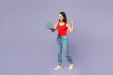 Full body young IT woman of Asian ethnicity wear casual clothes red tank shirt hold use work on laptop pc computer isolated on plain pastel light purple background studio portrait. Lifestyle concept.