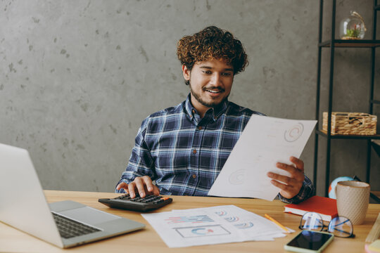 Successful Smart Smiling Young Employee Business Indian Man He Wears Casual Blue Checkered Shirt Read Paper Account Documents Using Calculator Sit Work At Office Desk With Laptop Pc Computer Indoors.