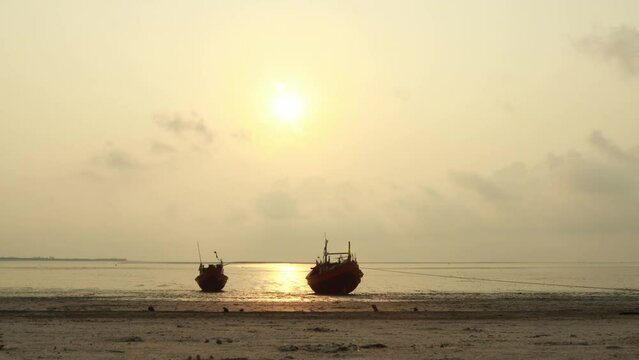 A time-lapse video of two small wooden fishing boats anchored on a calm sandy shore during a peaceful golden sunrise. The sun gently rises through soft hazy clouds, reflecting warm light 