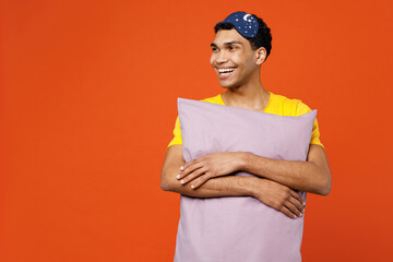 Calm young smiling happy fun man wearing pyjamas jam sleep eye mask rest relax at home hold pillow look aside on area isolated on plain orange background studio portrait. Good mood night nap concept.