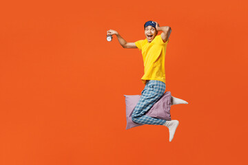 Full body side view young man wearing pyjamas jam sleep eye mask rest relax at home flying on pillow pov super hero hold clock isolated on plain orange background studio. Good mood night nap concept.