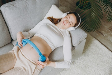 Young pregnant woman wear casual clothes put headphones on belly listen music lying on grey sofa...