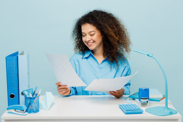 Young smiling hapy cheerful successful employee business woman wear casual shirt sit work at white office desk hold paper account documents isolated on plain pastel blue background studio portrait.
