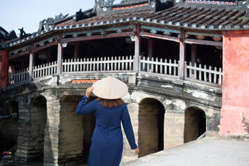 Woman traveler wearing Ao Dai Vietnamese dress sightseeing at Japanese covered bridge in Hoi An town, Vietnam. 