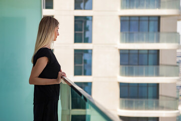 Side view of young cute woman in black on skyscraper balcony with view of Dubai UAE, thinking looking away. Pretty lady posing on terrace of tower block. Leisure activity concept. Copy ad text space