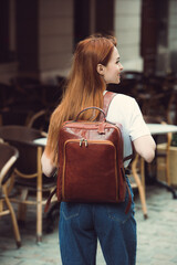 Naklejka na ściany i meble beautiful red-haired girl with red lips posing with a leather backpack. wearing blue jeans and a sweater