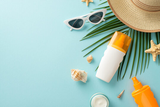 UV Safety Awareness Month. High angle view photo of sun protection lotion, sunglasses, straw hat and marine shells with palm leaf on light blue isolated background with copy-space