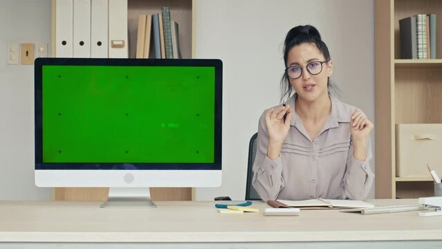 Medium shot of woman looking at camera and presenting application on monitor with green chroma key screen in office
