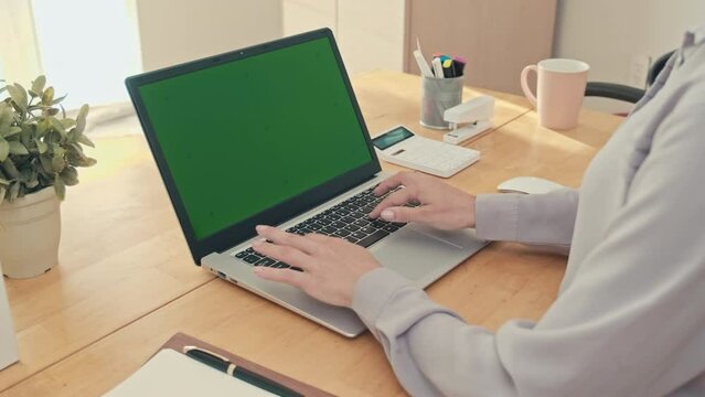 Over the shoulder shot of woman typing on keyboard of laptop with green chroma key screen at desk in office, slow motion