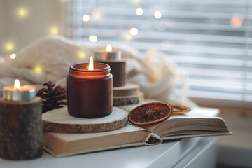 Cozy autumn or winter composition with aromatic candle, wool sweater, fairy lights, book. Aromatherapy, home atmosphere of cosiness and relax. Wooden background close up
