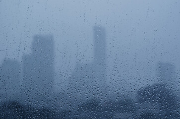 Rain drop on glass window in monsoon season with blurred city buildings background. - obrazy, fototapety, plakaty