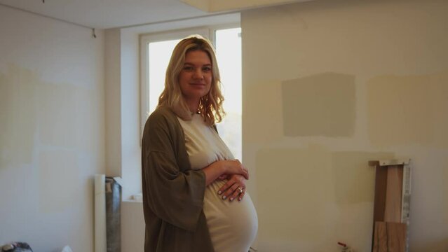 Beautiful caucasian pregnant woman standing in unfinished room, looking around. Optimistic future mother smiling into the camera. Visiting new home, relocating to bigger house for baby