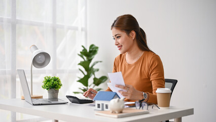 A beautiful Asian woman sits at her desk, paying her bills online on her laptop.