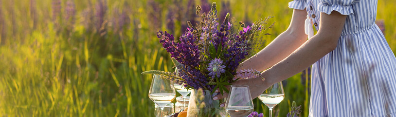 Beautiful happy young woman on a meadow arranging table for outdoor event, gathering wildflowers, lighting candles. Wedding or romantic date decoration in the field with purple lupins, fruits banner