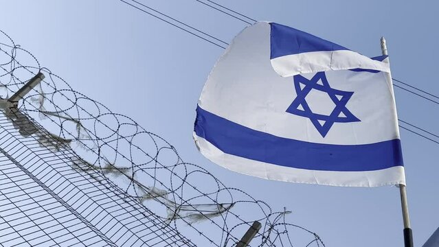 Israeli flag white and blue with star Magen David fluttering in wind on background blue sky and of barbed wire. outdoors. 