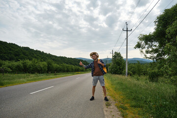 Hipster with hat and yellow backpack tries to stop car with thumbs up. Concept of traveling alone. Young Happy Caucasian man hitchhiking standing by road on sunny summer day.