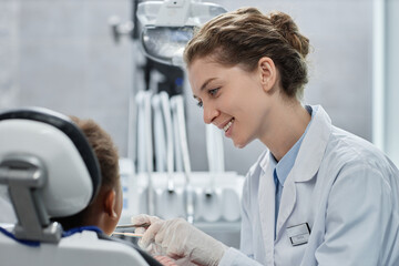 Side view portrait of smiling young woman as friendly female dentist working with child in dental...
