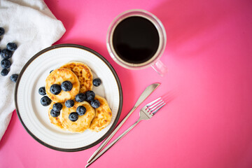 Top view, flat lay, Traditional cuisine of Russia and Ukraine. Cottage cheese pancakes on a white plate, a fork, coffee.Syrniki with blueberries on a pink background, top view. Sweet food 