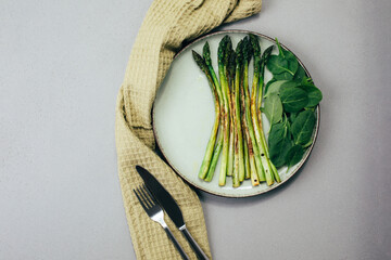 Cooked green asparagus on a green plate with fresh spinach leaves on a gray table with a green...
