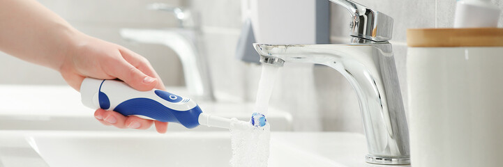 Woman washing electric toothbrush under flowing water in bathroom, closeup. Banner design