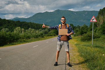 Guy went on trip alone, trying to stop passing car. Travel concept. Young man hitchhiker with backpack, stands by mountain country road and holds cardboard sign on sunny summer day. Front view.