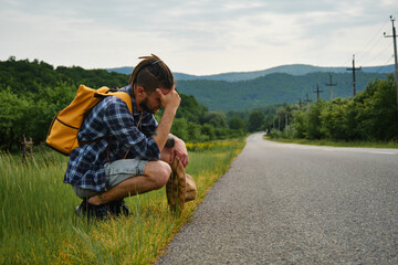 Naklejka na ściany i meble Guy trying to stop car. Hopelessness and despair. Young Caucasian man with dreadlocks and beard hitchhiking with backpack, sitting on side of empty country road and sad. Concept of traveling alone.