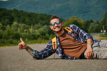 Young Caucasian man with dreadlocks and beard, hitchhiking in sunglasses and with backpack, lies in the middle of road in mountains. Travel concept. Guy trying to stop car with thumbs up.