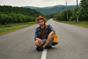 Travel concept. Guy tries to stop the car. Caucasian man hitchhiker with hat and backpack sitting with cardboard sign in middle of road on summer day.
