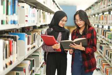 Asian young woman and muslim woman in hijab standing between book shelves, reading a book together...